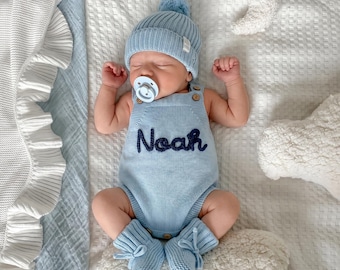 May include: A light blue knitted baby romper featuring the name Noah embroidered in dark blue thread across the chest, paired with a matching pom-pom beanie and booties. The outfit includes wooden buttons at the shoulders and a light blue pacifier.