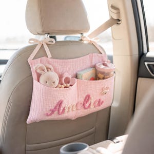 May include: A light pink muslin car seat organizer featuring two pockets, tied to the headrest with ribbons. The name Amelia is embroidered in pink script on the front. The pockets contain a plush rabbit, a teething ring, a book, and a cup.