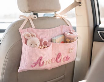 Peut inclure: Un organisateur de siège auto en mousseline rose clair avec deux poches, attaché à l'appui-tête par des rubans. Le prénom Amelia est brodé en lettres cursives roses sur le devant. Les poches contiennent un lapin en peluche, un anneau de dentition, un livre et un gobelet.