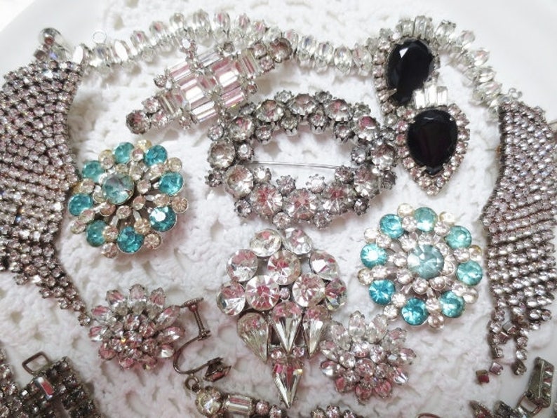 May include: A collection of vintage jewelry featuring a variety of styles, including crystal brooches, rhinestone necklaces, and a pair of black sunglasses.
