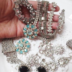 May include: A collection of vintage rhinestone and faux pearl brooches in silver and blue tones. The brooches feature various designs, including floral, geometric, and abstract patterns.