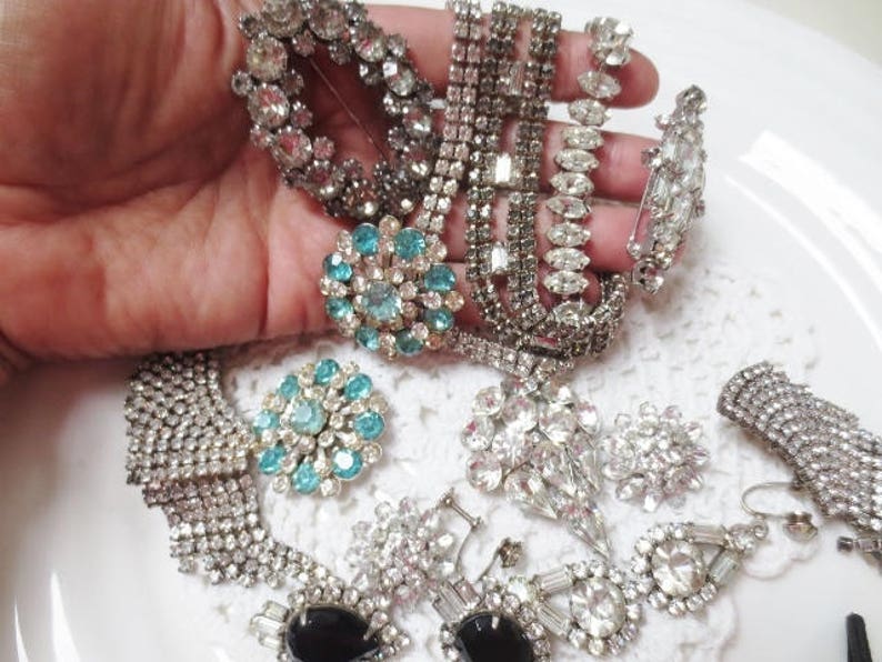 May include: A collection of vintage rhinestone and faux pearl brooches in silver and blue tones. The brooches feature various designs, including floral, geometric, and abstract patterns.