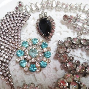 May include: A collection of vintage rhinestone brooches featuring a variety of designs, including a large blue and green floral brooch, a smaller brooch with a black and white geometric design, and a long, cascading brooch with a fringe of rhinestones.