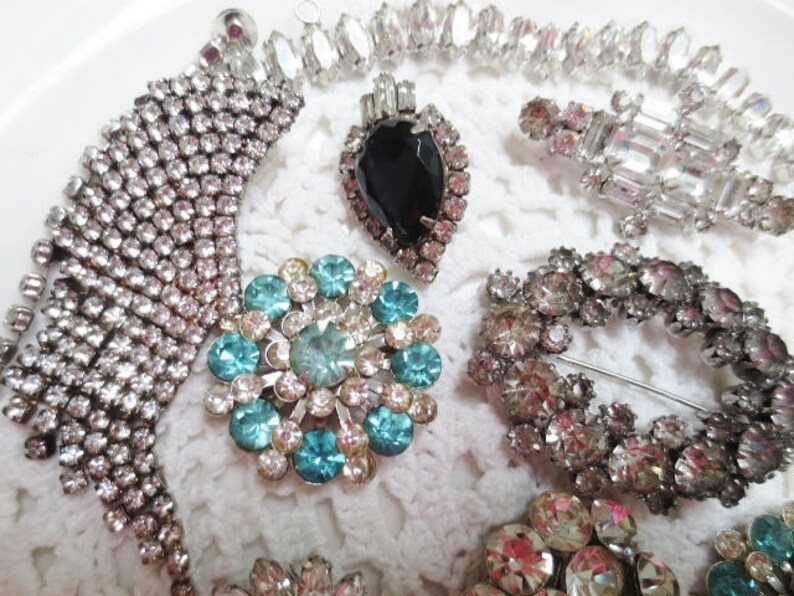 May include: A collection of vintage rhinestone brooches featuring a variety of designs, including a large blue and green floral brooch, a smaller brooch with a black and white geometric design, and a long, cascading brooch with a fringe of rhinestones.