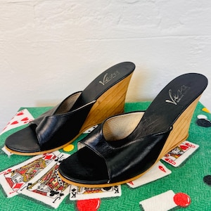 May include: Black leather wedge sandals with wooden heels. The sandals feature a strap across the toes and the brand name "Vera" on the insole. The heels are approximately 7.6 cm high. The sandals are displayed on a green surface.
