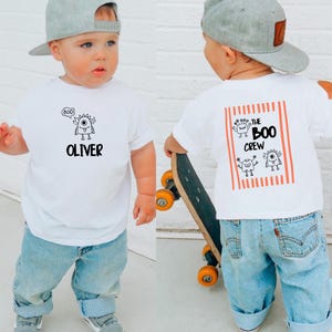 Könnte beinhalten: Ein weißes T-Shirt mit einer schwarzen Strichzeichnung eines Monsters, dem Text "BOO" und dem Namen "OLIVER" auf der Vorderseite sowie einem Rückendesign mit drei Monstern, dem Text "THE BOO CREW" und orangefarbenen vertikalen Streifen.