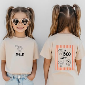 May include: A natural-coloured short-sleeve t-shirt featuring a small black monster graphic and the name AMELIA on the front. The back displays a rectangular orange striped design with three monster illustrations and the text THE BOO CREW.