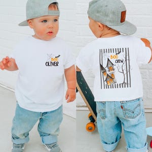 Könnte beinhalten: Ein weißes Kurzarm-T-Shirt mit einer kleinen Grafik auf der Brust, die ein Spinnennetz, eine Fledermaus, das Wort "BOO" in Orange und den Namen "OLIVER" in Schwarz zeigt. Auf der Rückseite befindet sich ein großes Motiv mit Spukhaus und "BOO CREW".