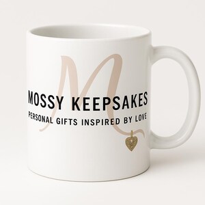 Könnte beinhalten: Weißer Keramikbecher mit gebogenem Henkel. Der Becher trägt den schwarzen Schriftzug "MOSSY KEEPSAKES" mit einem großen, hellbraunen "M" dahinter. Darunter steht "PERSONAL GIFTS INSPIRED BY LOVE". Ein goldener Herzanhänger hängt darunter.