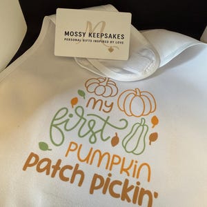 Könnte beinhalten: Weißes Baby-Lätzchen mit dem Text "my first pumpkin patch pickin'" in Grün und Orange. Das Lätzchen hat einen Klettverschluss und eine Geschenkkarte von Mossy Keepsakes.