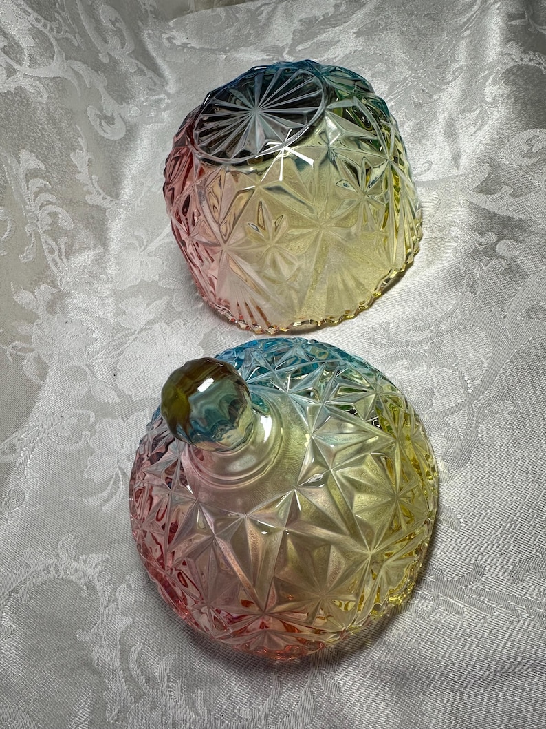 May include: Two decorative glass jars with lids. The jars are a rainbow of colors, including pink, yellow, and blue. The jars have a geometric starburst pattern. The lid of one jar is off, revealing the inside.