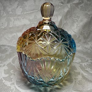 May include: Iridescent glass candy dish with a lid. The dish has a round shape with a diamond pattern and a decorative rim. The colors shift from yellow to blue and pink. The lid has a small, rounded handle.