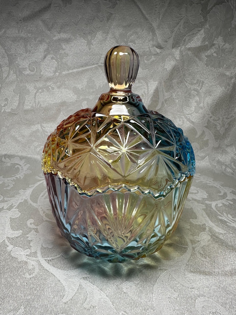 May include: Iridescent glass candy dish with a lid. The dish has a round shape with a diamond pattern and a decorative rim. The colors shift from yellow to blue and pink. The lid has a small, rounded handle.