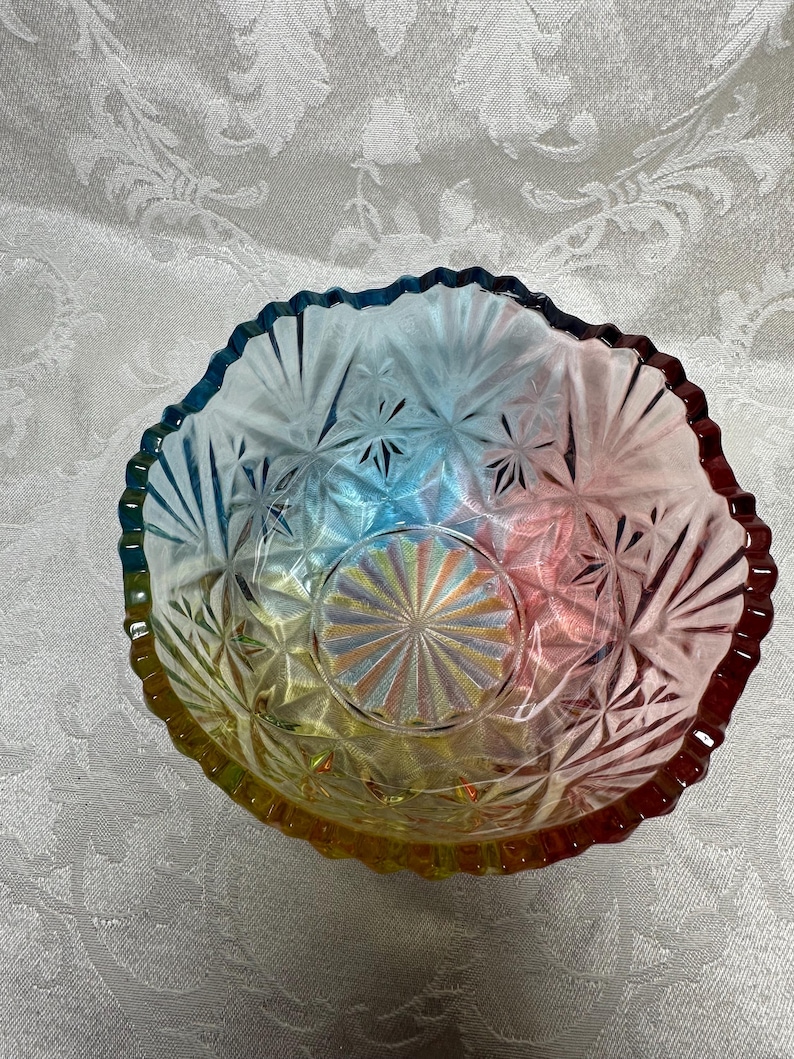 May include: A colorful, iridescent glass bowl with a starburst pattern. The bowl has a scalloped edge and transitions from yellow to red, blue, and green. The bowl is likely vintage and suitable for serving.