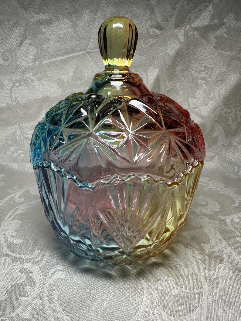 May include: A decorative glass jar with a lid. The jar is a mix of colors, including blue, pink, yellow, and red. The jar has a geometric pattern and a rounded shape. The lid has a rounded handle. The jar is likely used for storage or display.