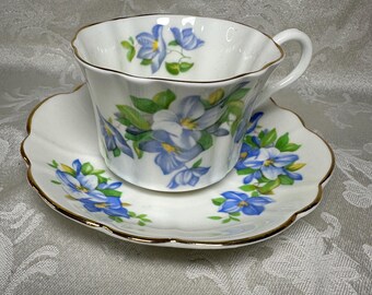 May include: A white teacup and saucer set with a floral design. The cup and saucer feature blue flowers with green leaves and gold trim. The cup has a scalloped rim, and the saucer has a matching shape.