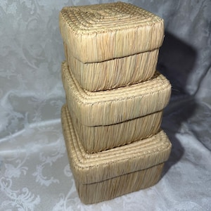 May include: A stack of three square woven boxes in varying sizes. The boxes are made of a light tan, straw-like material. The lids and sides are neatly woven, creating a textured surface. The boxes are stacked on top of each other.