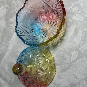 May include: A colorful, iridescent glass bowl and lid set. The bowl and lid are a mix of red, yellow, blue, and green hues. The bowl has a scalloped edge and a starburst pattern. The lid has a round knob.
