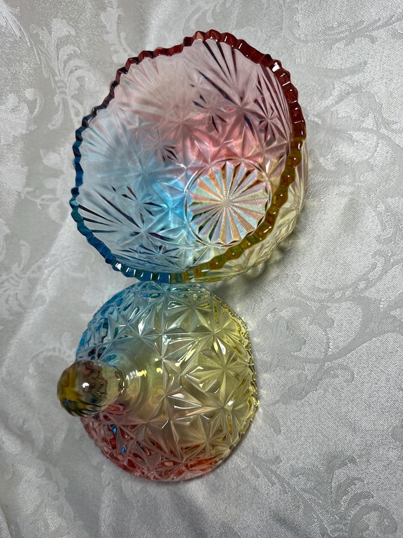 May include: A colorful, iridescent glass bowl and lid set. The bowl and lid are a mix of red, yellow, blue, and green hues. The bowl has a scalloped edge and a starburst pattern. The lid has a round knob.