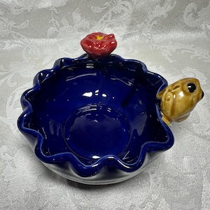 Puede incluir: Un cuenco de cerámica con forma de tortuga, con interior azul oscuro y borde ondulado. Una pequeña flor roja se encuentra encima del cuenco, y una cabeza de tortuga marrón se extiende desde un lado. El cuenco es probablemente decorativo.