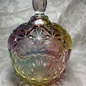 May include: A decorative glass candy dish with a lid. The dish is a mix of colors, including pink, yellow, and blue. The glass has a starburst pattern and a knob on the lid. The dish is approximately 6 inches tall.