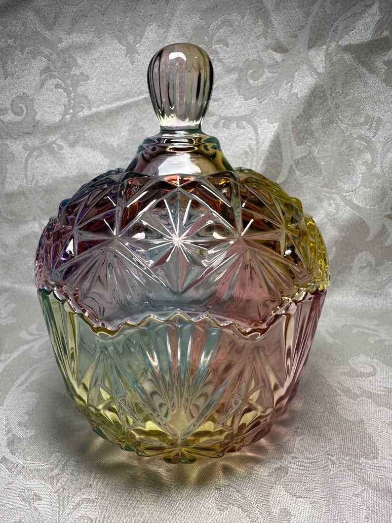 May include: A decorative glass candy dish with a lid. The dish is a mix of colors, including pink, yellow, and blue. The glass has a starburst pattern and a knob on the lid. The dish is approximately 6 inches tall.