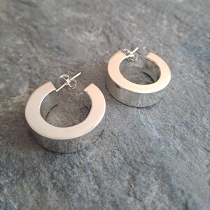May include: A pair of silver hoop earrings. The earrings are thick and have a rounded, rectangular shape. The earrings are displayed on a grey, textured surface.