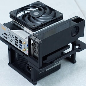 May include: A black computer case with a silver motherboard, a black fan, and a black graphics card. The components are stacked on top of each other, showcasing a compact computer build.
