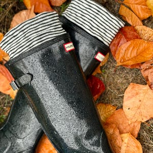 May include: Black Hunter wellington boots with water droplets, and striped socks. The boots feature a buckle and a red Hunter logo. Orange and brown autumn leaves are scattered around the boots.