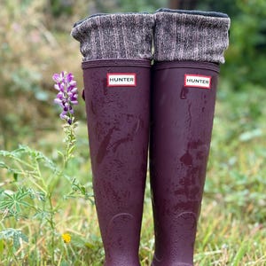 May include: Plum-coloured Hunter wellington boots with grey knit boot toppers. The boots feature the Hunter logo and are set in a grassy area with a purple flower. The boots are designed for wet weather.