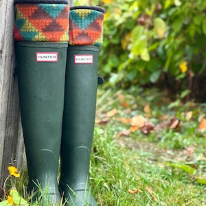 May include: Dark green Hunter wellington boots with patterned boot toppers. The boots are in a grassy area next to a wooden post. The Hunter logo is displayed on the front of each boot.