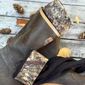 May include: Brown rubber boots with tan soles and a camouflage patterned lining. The boots have a logo on the side and are resting on a weathered wooden surface. The boots are designed for outdoor use.