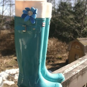 May include: A pair of teal blue rain boots with white fleece cuffs and a blue flower accent. The boots are on a wooden railing.