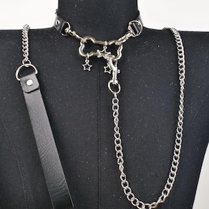 May include: A black leather choker with silver-toned hardware, a chain leash, and a matching handle. The choker features a decorative centerpiece with star charms. The chain and handle are attached with metal rings and clasps.
