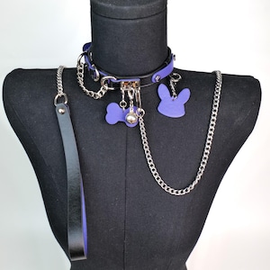May include: A purple and black pet collar set with a matching leash and chain. The collar features a bone-shaped tag, a bunny-shaped tag, and a small bell. The leash is black with a purple interior.