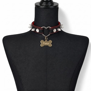 May include: A black leather choker with red lining, featuring silver-toned metal spikes and studs. A central silver heart-shaped ring holds a gold-toned bone-shaped tag engraved with the word Puppy in a serif font, centred on the collar band.