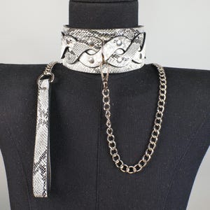 May include: A black and white snakeskin-patterned choker collar with a matching leash and silver chain. The wide collar features a braided design and silver-toned rivets. The leash has a handle and a silver clasp. The chain is attached to the collar.