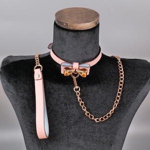 Könnte beinhalten: Ein rosa Haustierhalsband und Leinen-Set. Das Halsband hat eine Schleife mit goldfarbener Hardware und einem hellblauen Mittelteil. Eine goldfarbene Kette und eine passende rosa Leine vervollständigen das Set. Auf einer schwarzen Schaufensterpuppe ausgestellt.