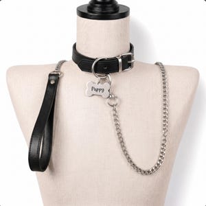 May include: A black leather collar with a silver-toned buckle and a bone-shaped metal tag engraved with the word Puppy. The collar features a silver metal chain leash and a black leather handle, all displayed on a neutral mannequin torso.