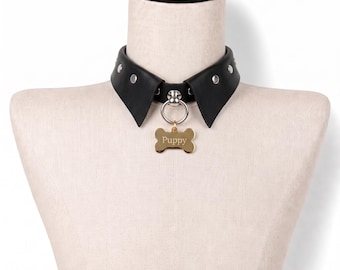 May include: A black collar featuring a pointed collar-style design with silver-toned metal studs along the band. A silver-toned metal O-ring is attached to the center, holding a gold-toned bone-shaped tag with the word Puppy engraved on it.