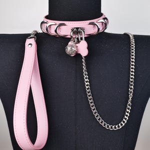 May include: A pink leather collar with silver-tone metal hardware, including rings and a clasp. A matching leash and a chain are attached. A pink bone-shaped charm and a silver bell dangle from the collar.