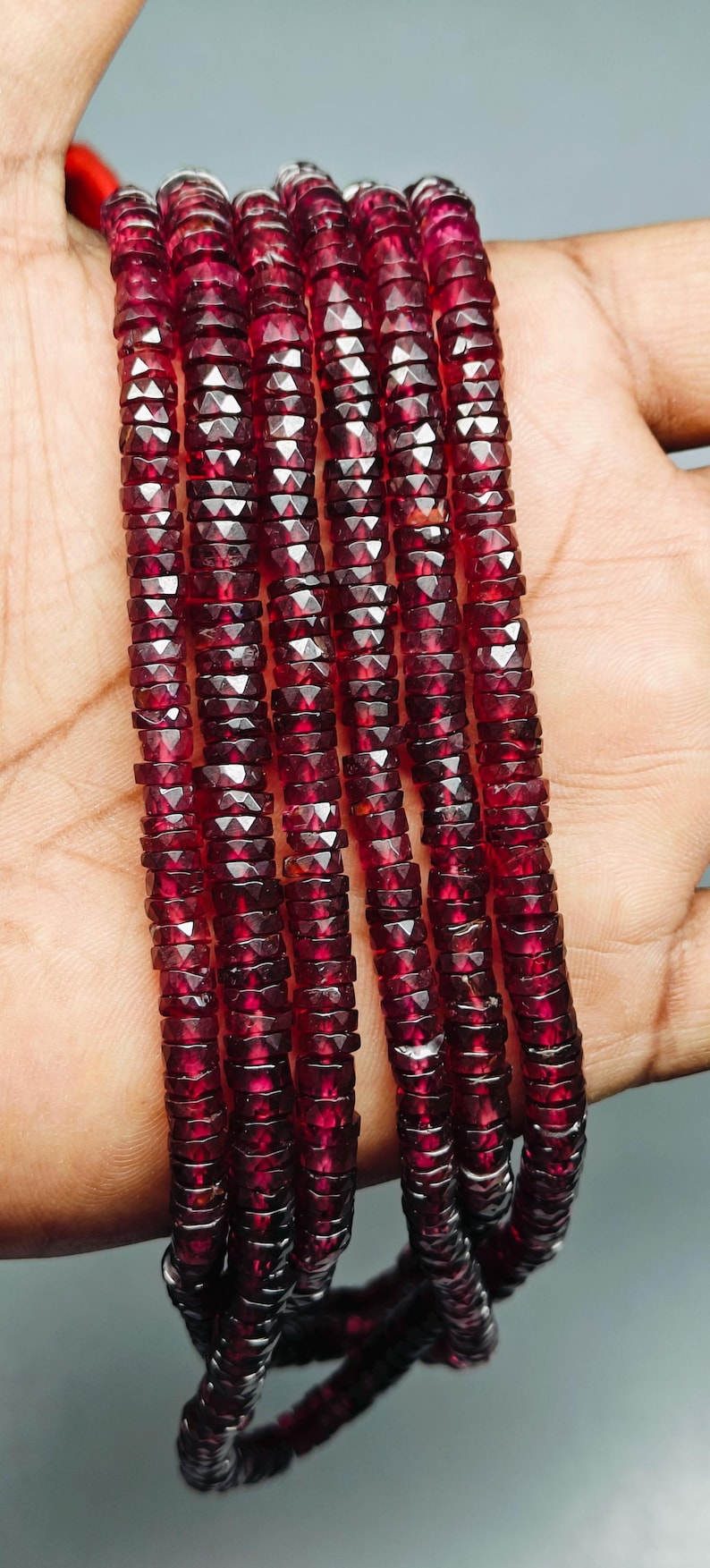 May include: Strands of faceted, deep red gemstones. The beads are small, rectangular, and strung together, creating a necklace or bracelet. The color is a rich, dark red, with a slight sparkle.