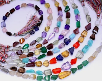 May include: Assortment of gemstone bead strands in various colors, including purple, brown, yellow, green, and blue. Each strand features faceted, irregular-shaped beads. Tassels with colorful threads are attached to the ends of some strands.