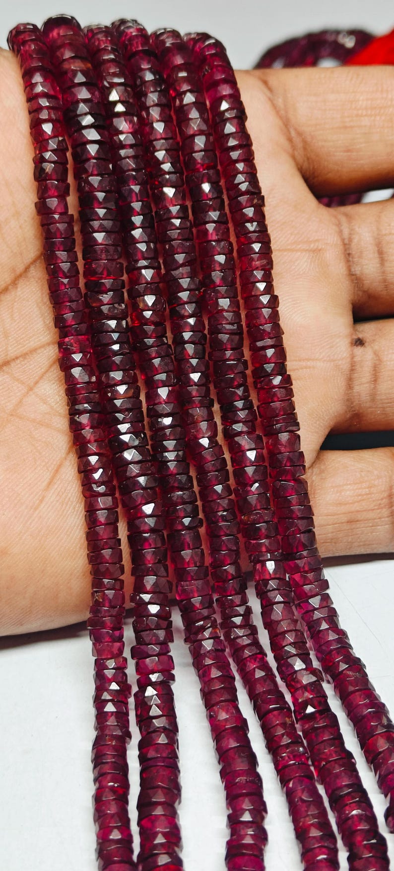 May include: Strands of faceted, deep red gemstone beads. The beads are small, flat, and disc-shaped, strung together to form multiple strands. The color is a rich, dark red, with a slight sparkle from the facets. The beads are held in a hand.