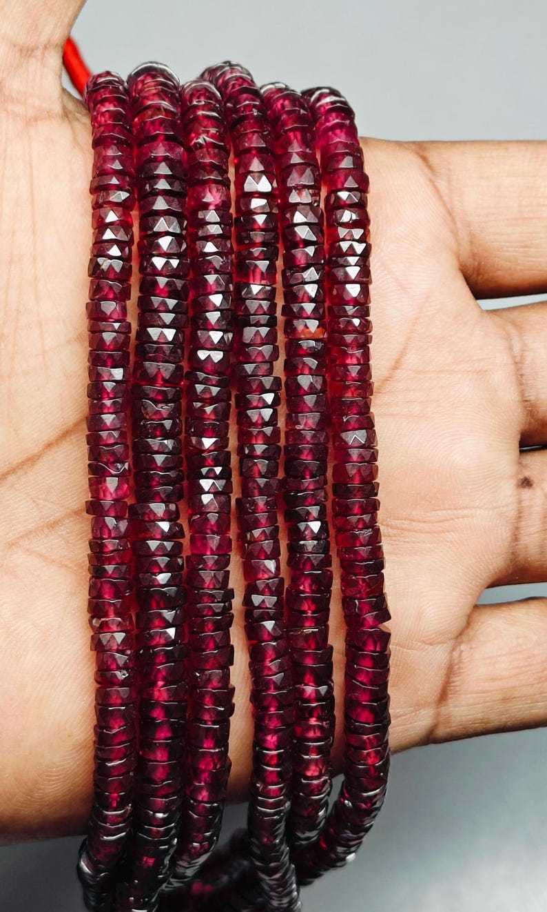 May include: Strands of faceted, deep red gemstone beads. The beads are small, with a geometric cut that catches the light, creating a sparkling effect. The beads are strung together, forming multiple strands. The overall color is a rich, dark red.