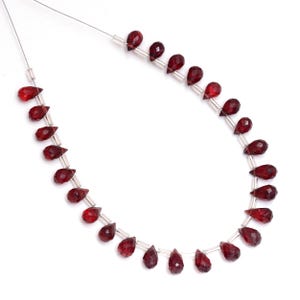 Puede incluir: Un collar de cuentas de granate rojo oscuro facetadas en forma de lágrima. Las cuentas están ensartadas en un fino alambre plateado. Las cuentas están espaciadas uniformemente y tienen una superficie pulida y reflectante.