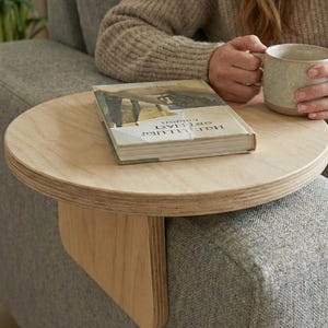 May include: A circular wooden side table in a light colour, with a book and a mug on top. The table is attached to the arm of a grey sofa. The book has an illustrated cover. The mug is pale green.