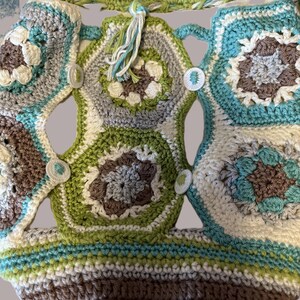 May include: A handmade crochet bag in shades of brown, green, blue, gray, and white. The bag features a patchwork design with floral and geometric patterns, connected by crocheted straps and secured with white buttons. The bag has a tassel.