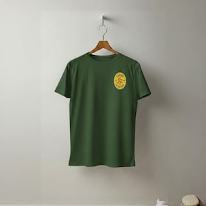May include: A green t-shirt with a yellow oval emblem on the left chest. The emblem features the words "Camino de Santiago" in a circular design with a stylized figure. The shirt is hanging on a wooden hanger.