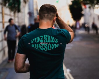 Pode incluir: Uma t-shirt verde-escura com o texto "FUCKING ASTURIAS IS AWESOME" numa fonte estilizada. A pessoa está a usar calças de ganga azuis e a andar numa rua da cidade. A imagem é tirada por trás.